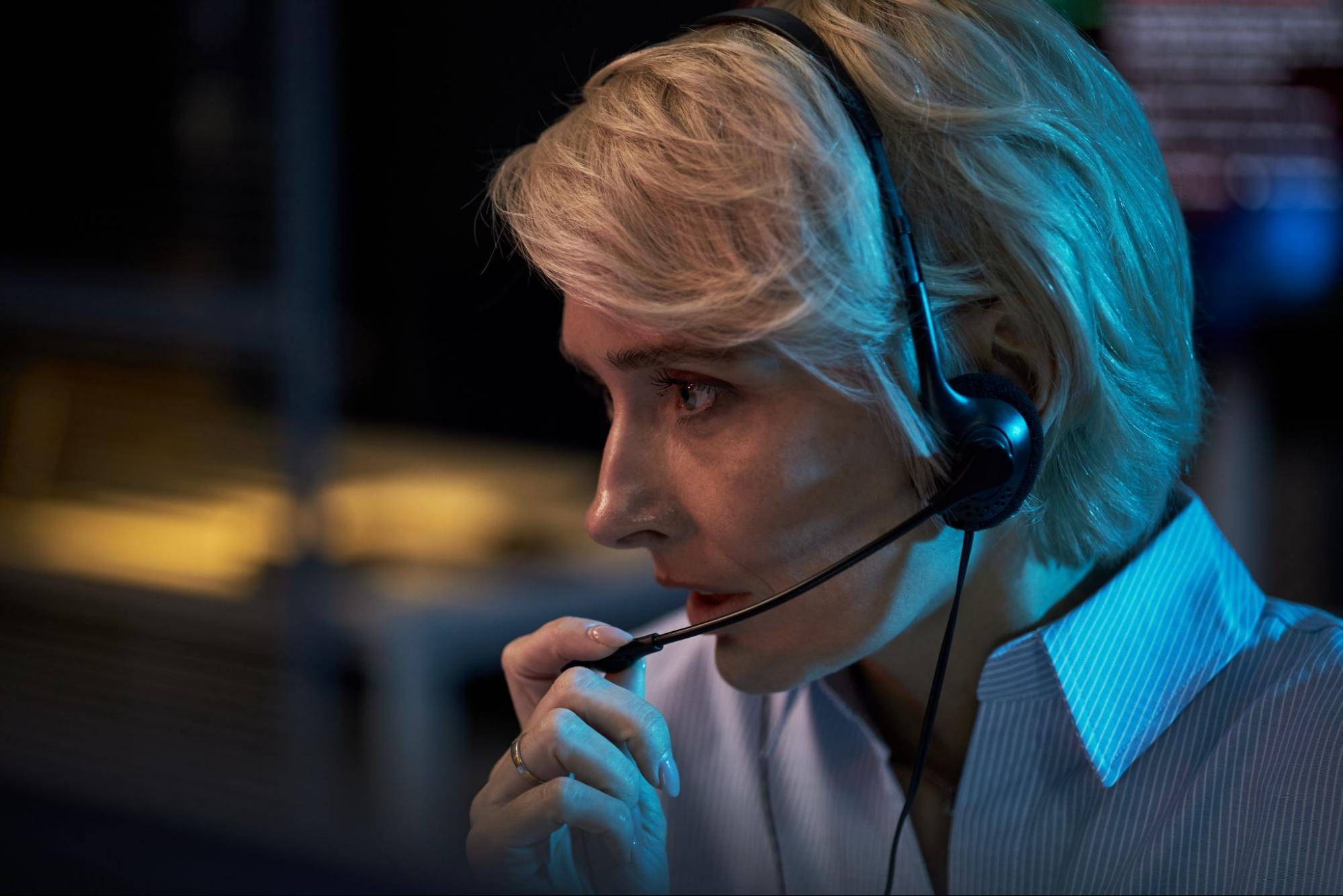 Close up of middle aged Caucasian woman wearing headset, speaking into microphone while monitoring screen in office.
