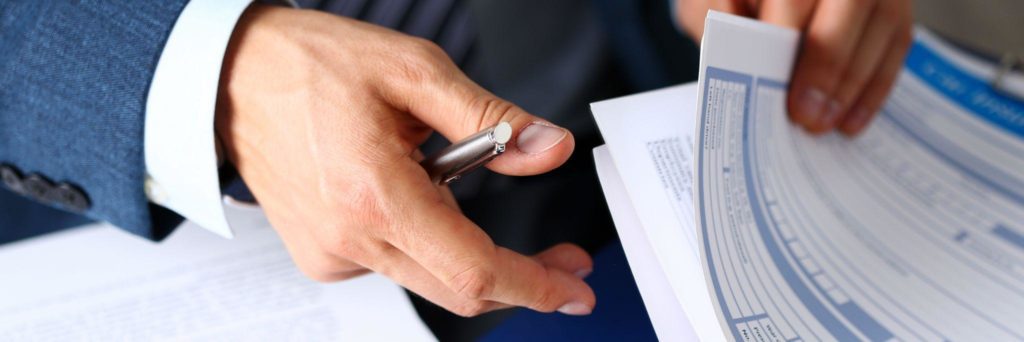 Male arm in suit offer insurance form clipped to pad and silver pen to sign closeup.