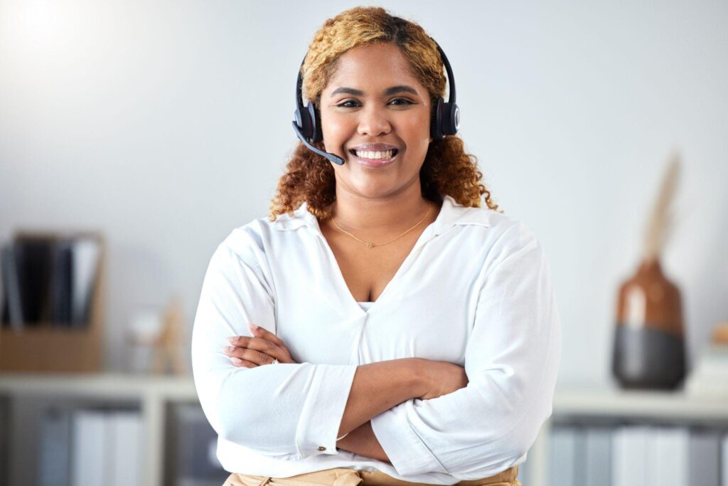 Female telephone answering receptionist with headset and white shirt with her arms crossed standing up.